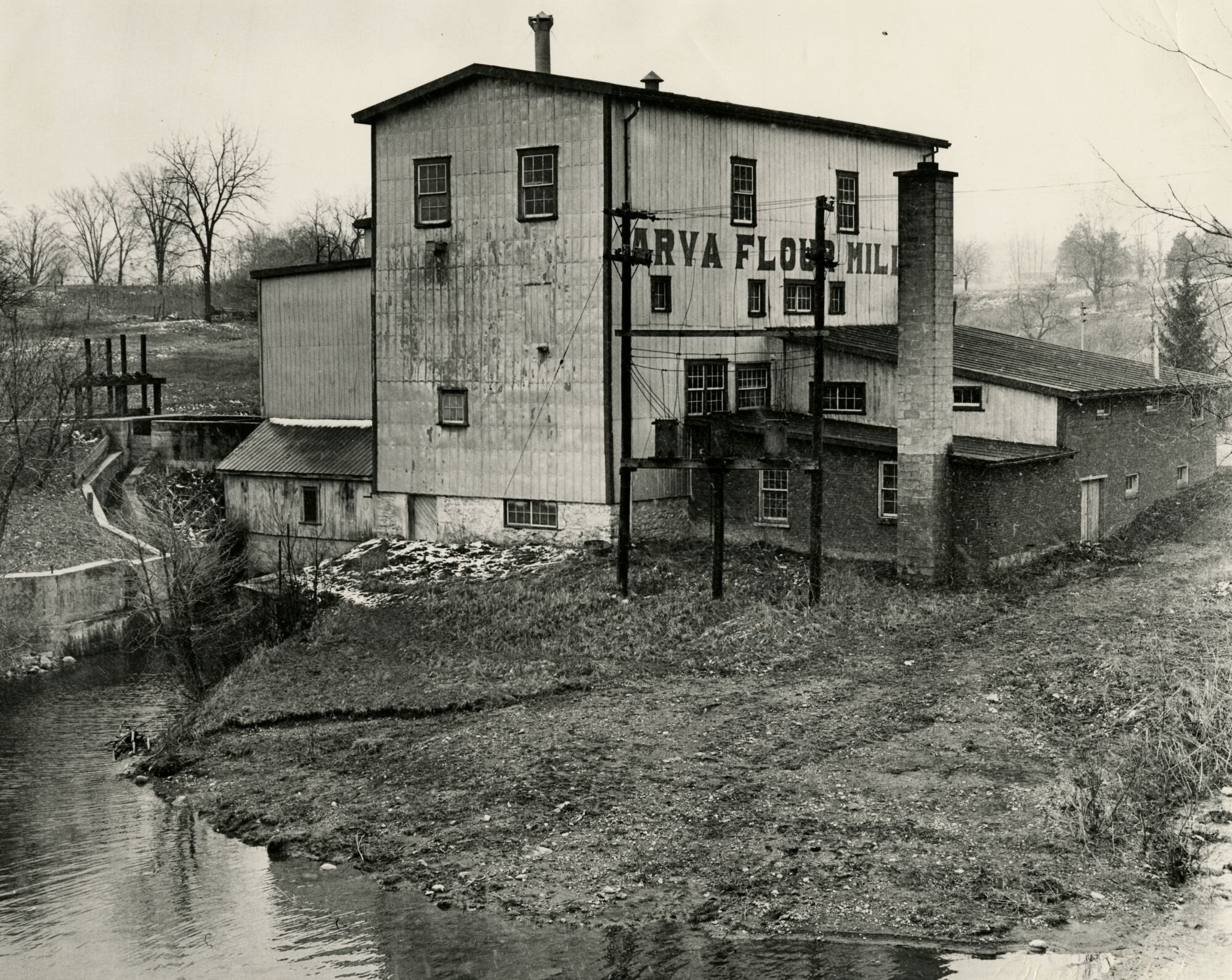 Early black-and-white photograph of the Arva Flour Mill on the banks of Medway Creek, with 'ARVA FLOUR MILLS' painted on the timber siding and the head-race visible along the water