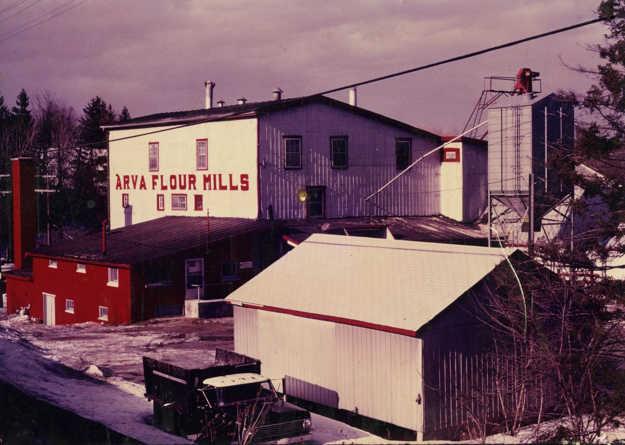 Colour photograph of the Arva Flour Mill in the 1970s or 1980s, with 'ARVA FLOUR MILLS' painted in red on the white timber siding and a grain elevator alongside a truck at the loading dock